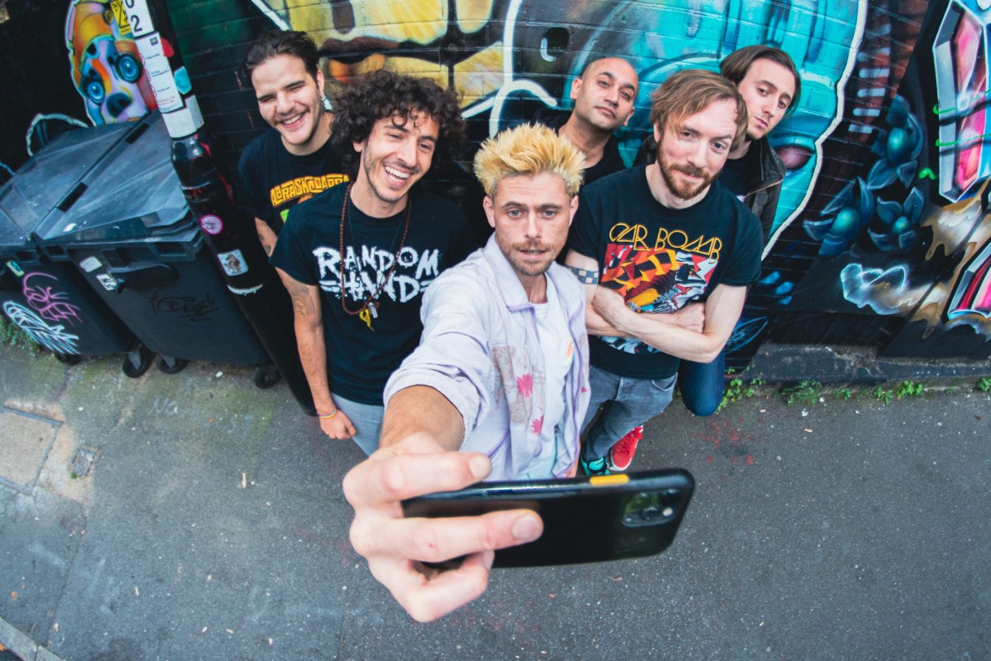 Six people stand close together in a narrow outdoor alley, posing for a group selfie taken from above with a wide‑angle lens. One person in the foreground holds a smartphone toward the camera, with the phone and outstretched arm dominating the lower center of the image. The group is positioned against a brick wall covered in brightly colored graffiti featuring abstract shapes and faces. Several people wear graphic T‑shirts, and one wears a light-colored overshirt. To the left, large black rubbish bins line the alley beside a sticker-covered pole. The ground is gray asphalt with small debris, and the scene has a casual, urban street setting with bold colors and strong perspective distortion.