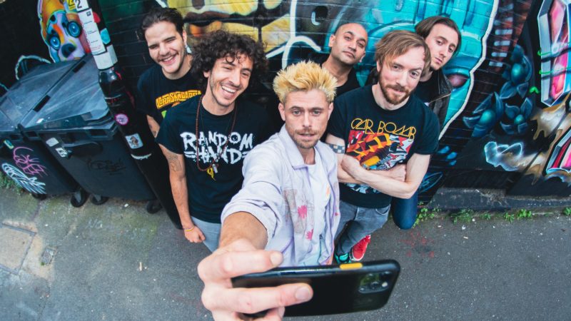 Six people stand close together in a narrow outdoor alley, posing for a group selfie taken from above with a wide‑angle lens. One person in the foreground holds a smartphone toward the camera, with the phone and outstretched arm dominating the lower center of the image. The group is positioned against a brick wall covered in brightly colored graffiti featuring abstract shapes and faces. Several people wear graphic T‑shirts, and one wears a light-colored overshirt. To the left, large black rubbish bins line the alley beside a sticker-covered pole. The ground is gray asphalt with small debris, and the scene has a casual, urban street setting with bold colors and strong perspective distortion.