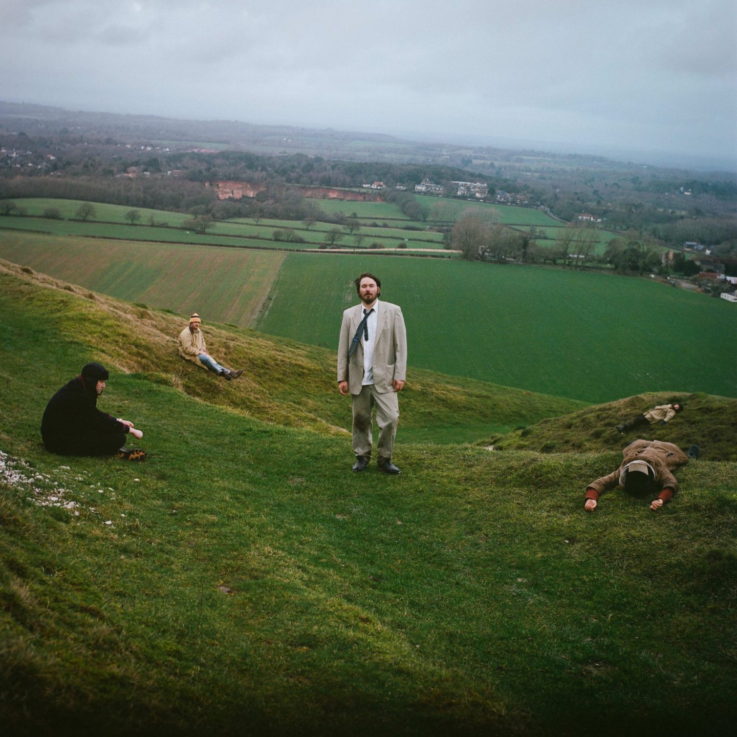 A moody outdoor portrait of a band spread across rolling green hills under an overcast sky. One person stands centrally in a light beige suit and loosened tie, facing the camera, while four others are positioned around the hillside — sitting or lying on the grass — creating a surreal, cinematic composition. The landscape stretches into the distance with patchwork fields, trees and misty countryside, giving the image a melancholic, atmospheric feel.
