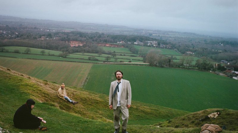 A moody outdoor portrait of a band spread across rolling green hills under an overcast sky. One person stands centrally in a light beige suit and loosened tie, facing the camera, while four others are positioned around the hillside — sitting or lying on the grass — creating a surreal, cinematic composition. The landscape stretches into the distance with patchwork fields, trees and misty countryside, giving the image a melancholic, atmospheric feel.