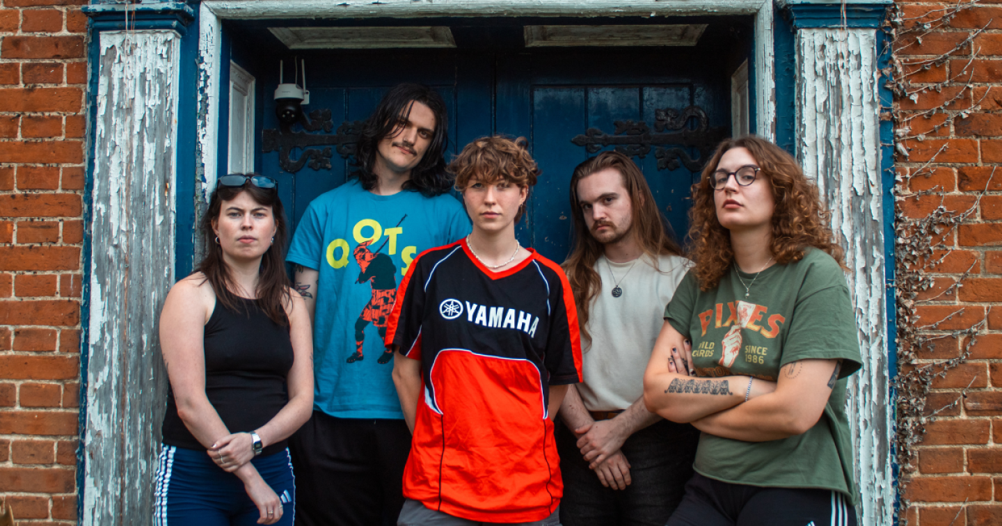 Five young adults standing together in front of a weathered blue doorway, posing seriously outdoors.
