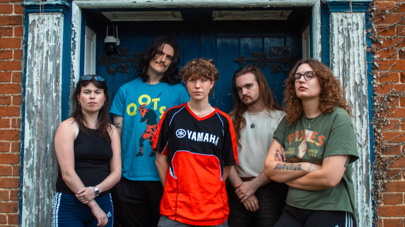 Five young adults standing together in front of a weathered blue doorway, posing seriously outdoors.