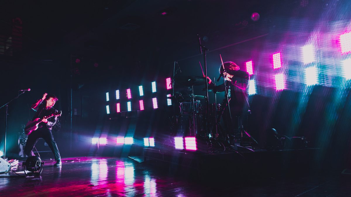 Live band performance on a dark stage, with one musician playing guitar and another at a drum kit, both lit by bright pink, blue and white stage lights that reflect across the glossy floor, creating a vivid, high-energy concert atmosphere.