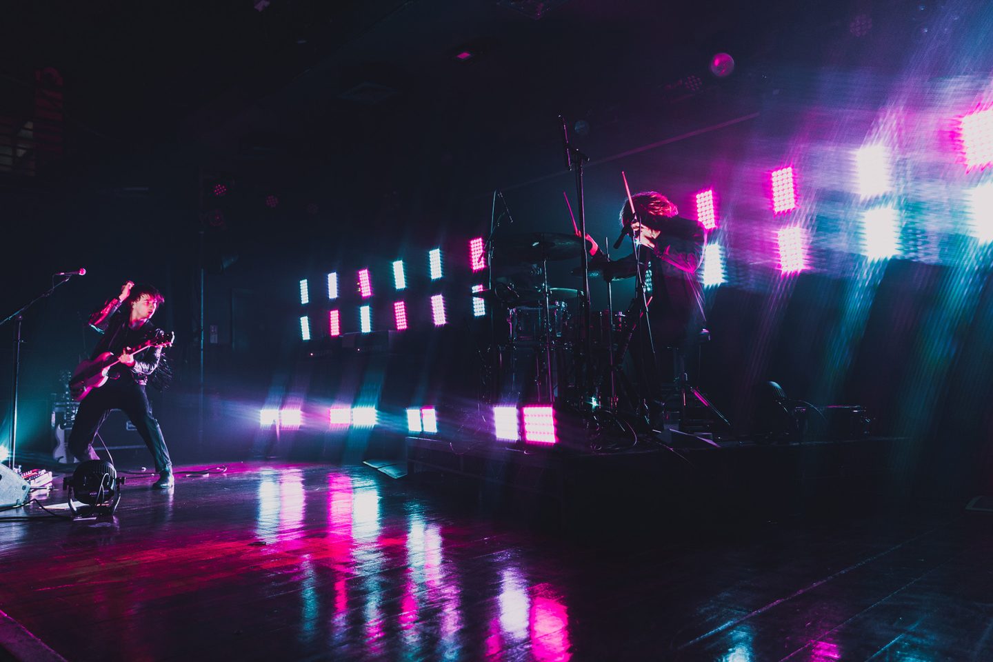 Live band performance on a dark stage, with one musician playing guitar and another at a drum kit, both lit by bright pink, blue and white stage lights that reflect across the glossy floor, creating a vivid, high-energy concert atmosphere.