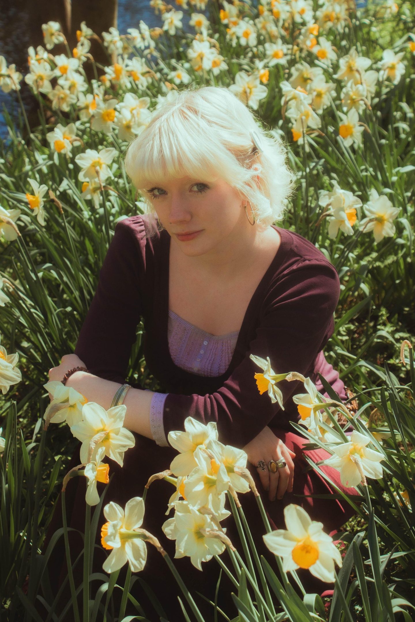 Person with short platinum-blonde hair sitting among blooming daffodils in soft sunlight.