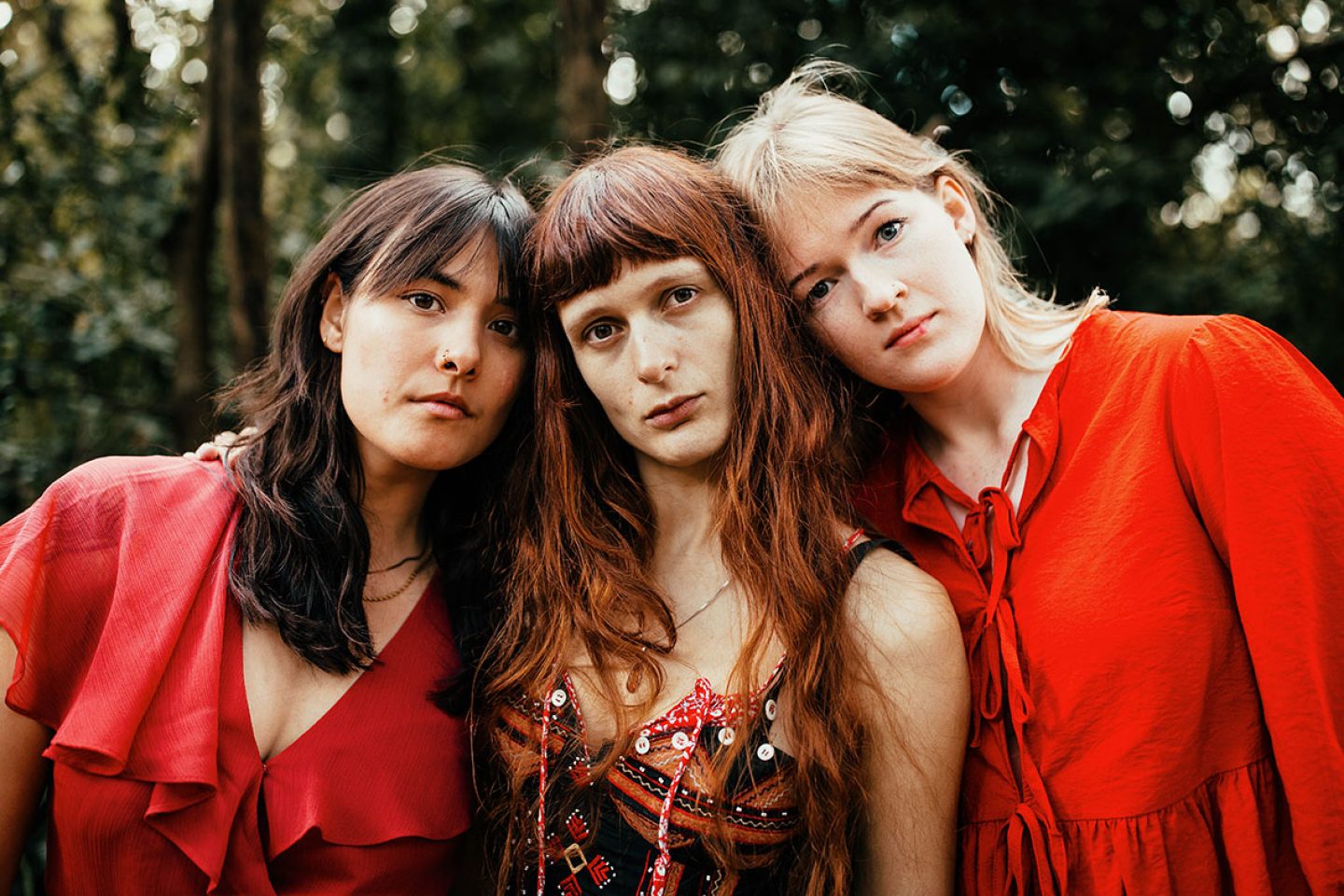 Three people stand closely together outdoors, facing the camera, wearing coordinated red clothing against a softly blurred backdrop of trees and greenery.