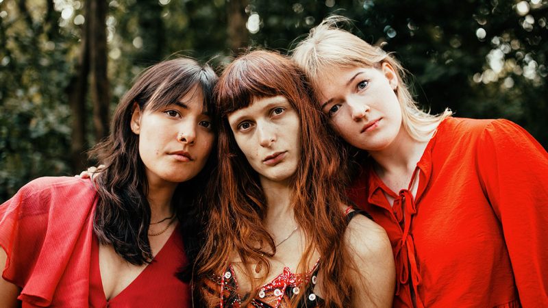 Three people stand closely together outdoors, facing the camera, wearing coordinated red clothing against a softly blurred backdrop of trees and greenery.