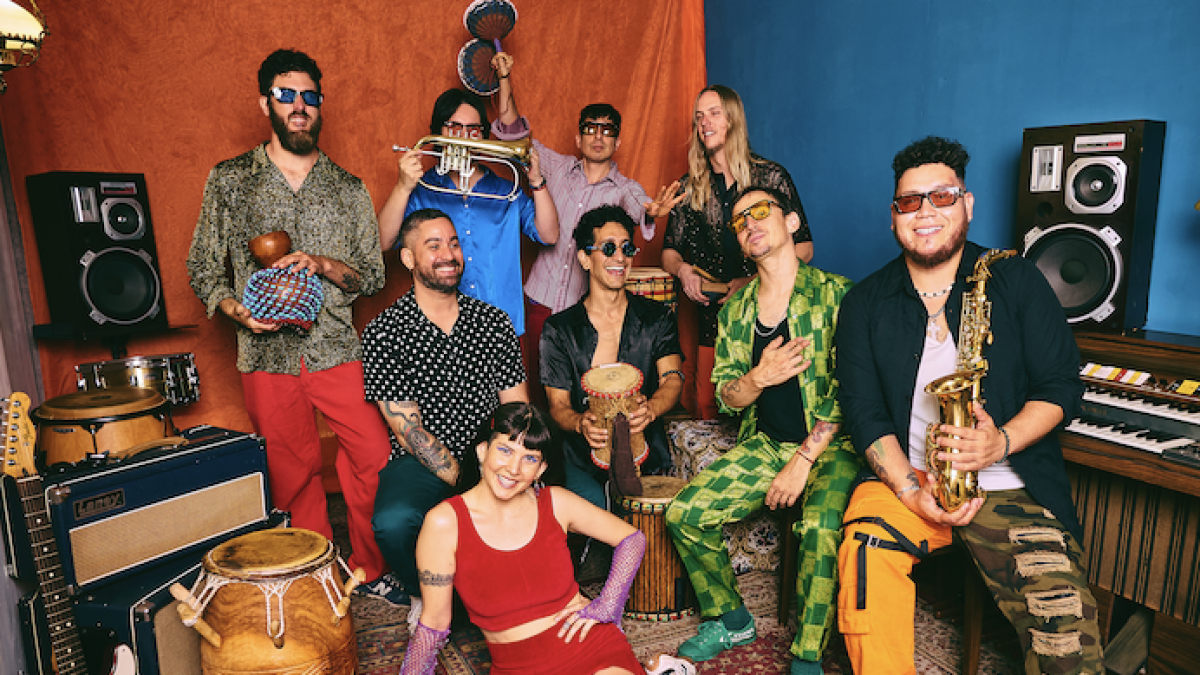 A colorful group portrait of a multicultural band posed in a music studio filled with instruments. Nine people wear bright, eclectic outfits and sunglasses while holding or sitting near percussion instruments, a saxophone, a trumpet, guitars, speakers, and a keyboard. A woman in a red outfit sits in the foreground on a patterned rug, while the others stand or sit behind her against bold orange and blue backdrops, creating a lively, energetic atmosphere.
