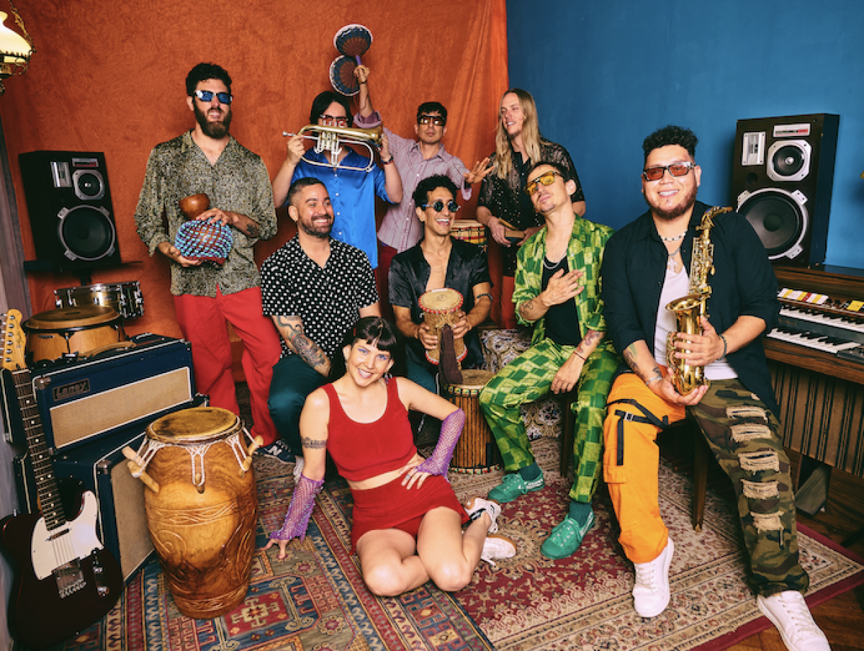 A colorful group portrait of a multicultural band posed in a music studio filled with instruments. Nine people wear bright, eclectic outfits and sunglasses while holding or sitting near percussion instruments, a saxophone, a trumpet, guitars, speakers, and a keyboard. A woman in a red outfit sits in the foreground on a patterned rug, while the others stand or sit behind her against bold orange and blue backdrops, creating a lively, energetic atmosphere.
