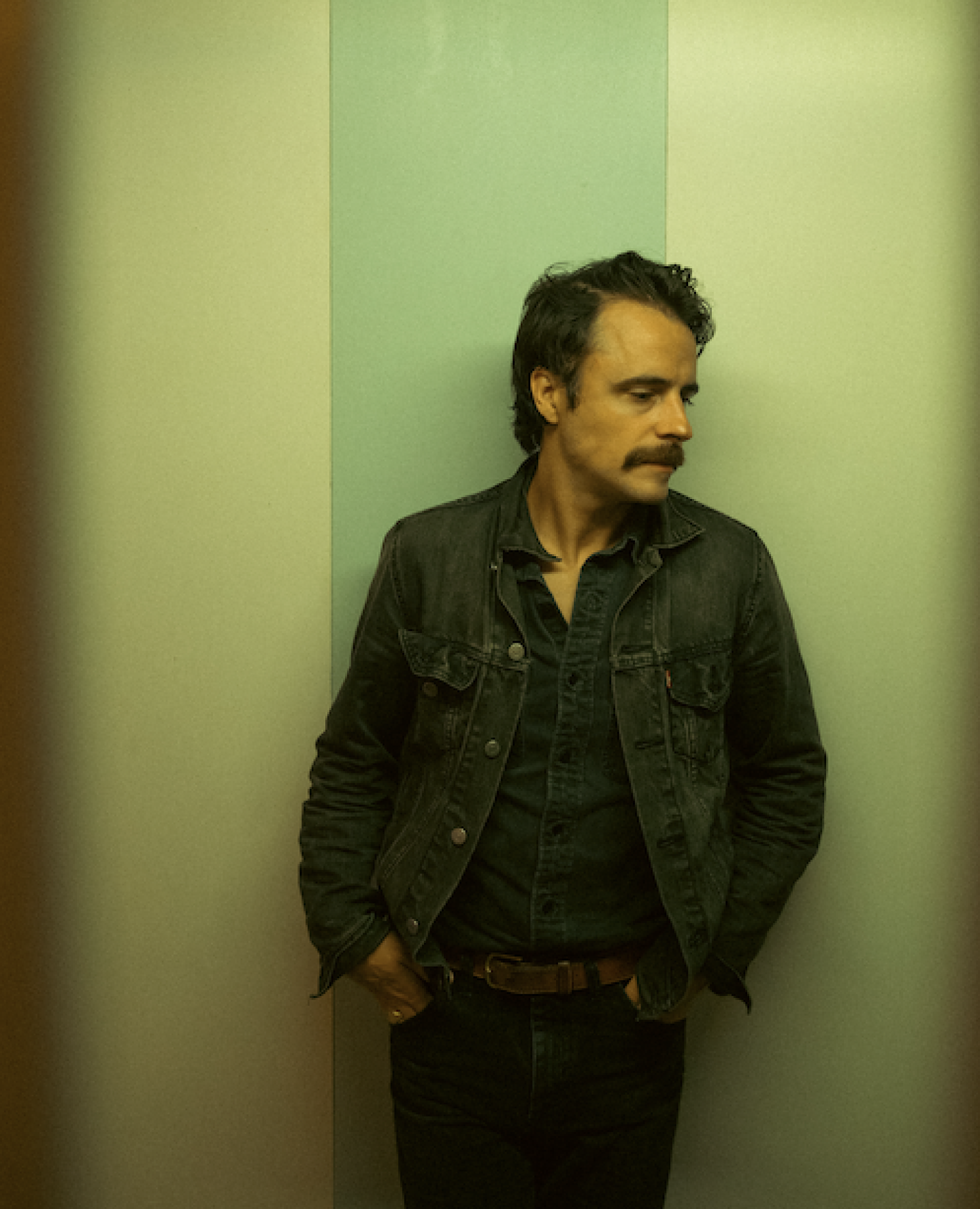 A moody portrait of a man standing against a vertically striped wall in muted green and beige tones. He’s wearing a dark denim jacket over a button-up shirt and jeans, with his hands in his pockets and his head turned to the side. The lighting and slight vignette give the image a cinematic, vintage feel.
