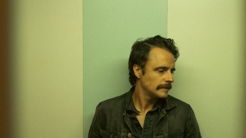 A moody portrait of a man standing against a vertically striped wall in muted green and beige tones. He’s wearing a dark denim jacket over a button-up shirt and jeans, with his hands in his pockets and his head turned to the side. The lighting and slight vignette give the image a cinematic, vintage feel.