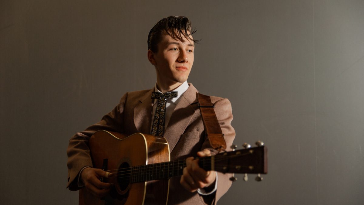A young man in a vintage-style suit plays an acoustic guitar against a plain background.