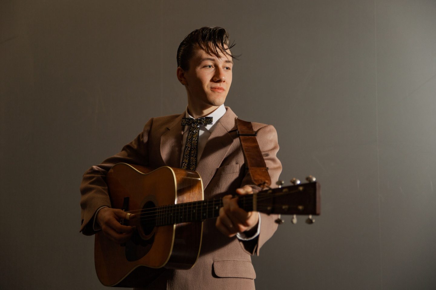 A young man in a vintage-style suit plays an acoustic guitar against a plain background.