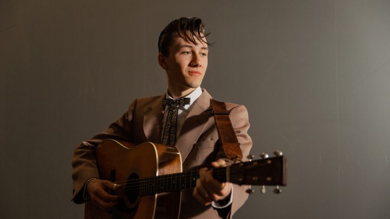 A young man in a vintage-style suit plays an acoustic guitar against a plain background.