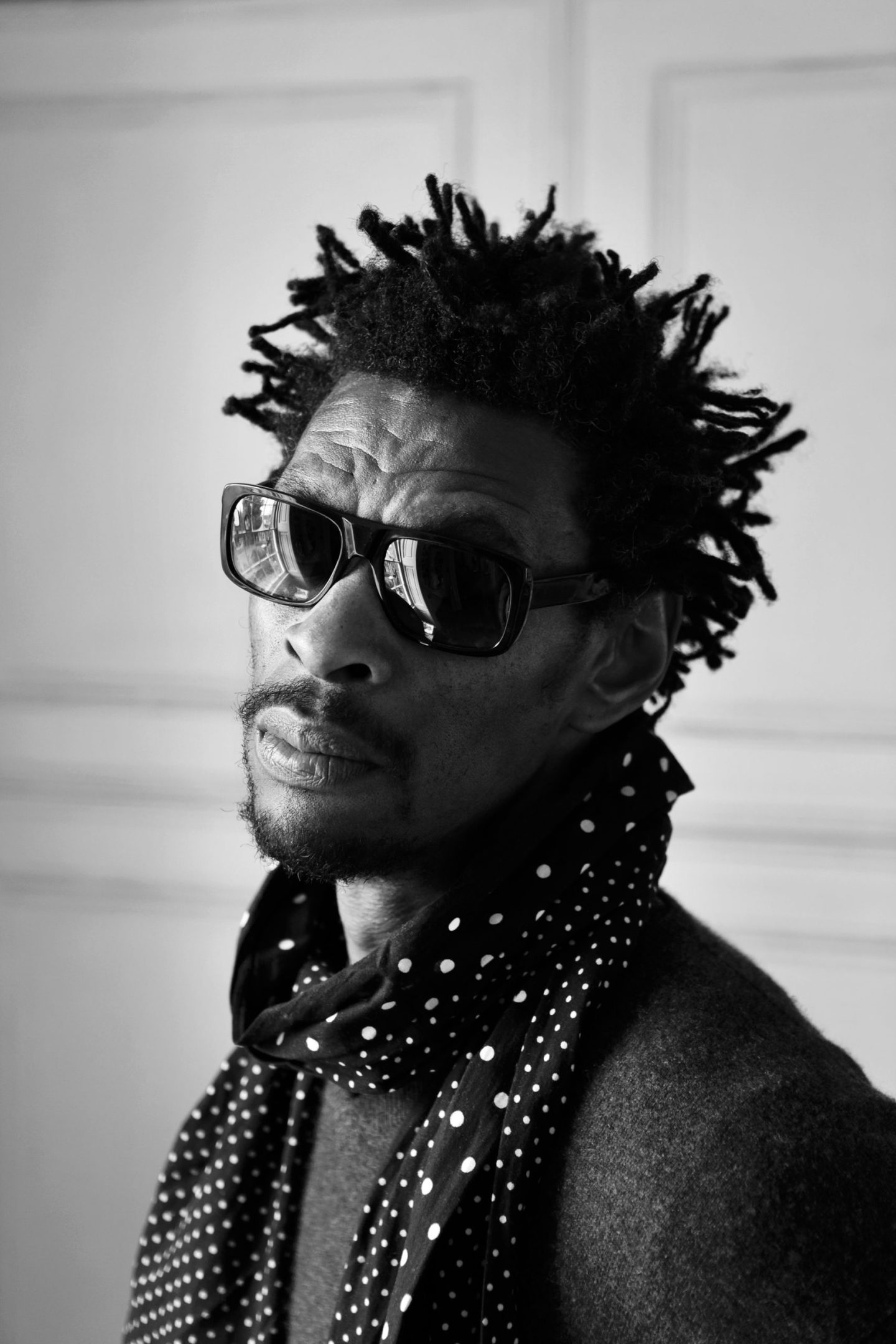 Black-and-white portrait of a person wearing sunglasses and a polka-dot scarf, looking over their shoulder.