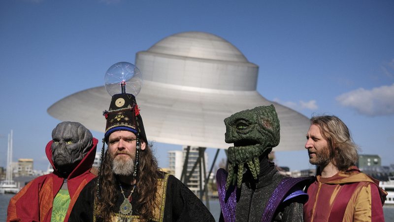 Four costumed people dressed as alien and fantasy characters stand by a waterfront, with a UFO-shaped structure in the background.