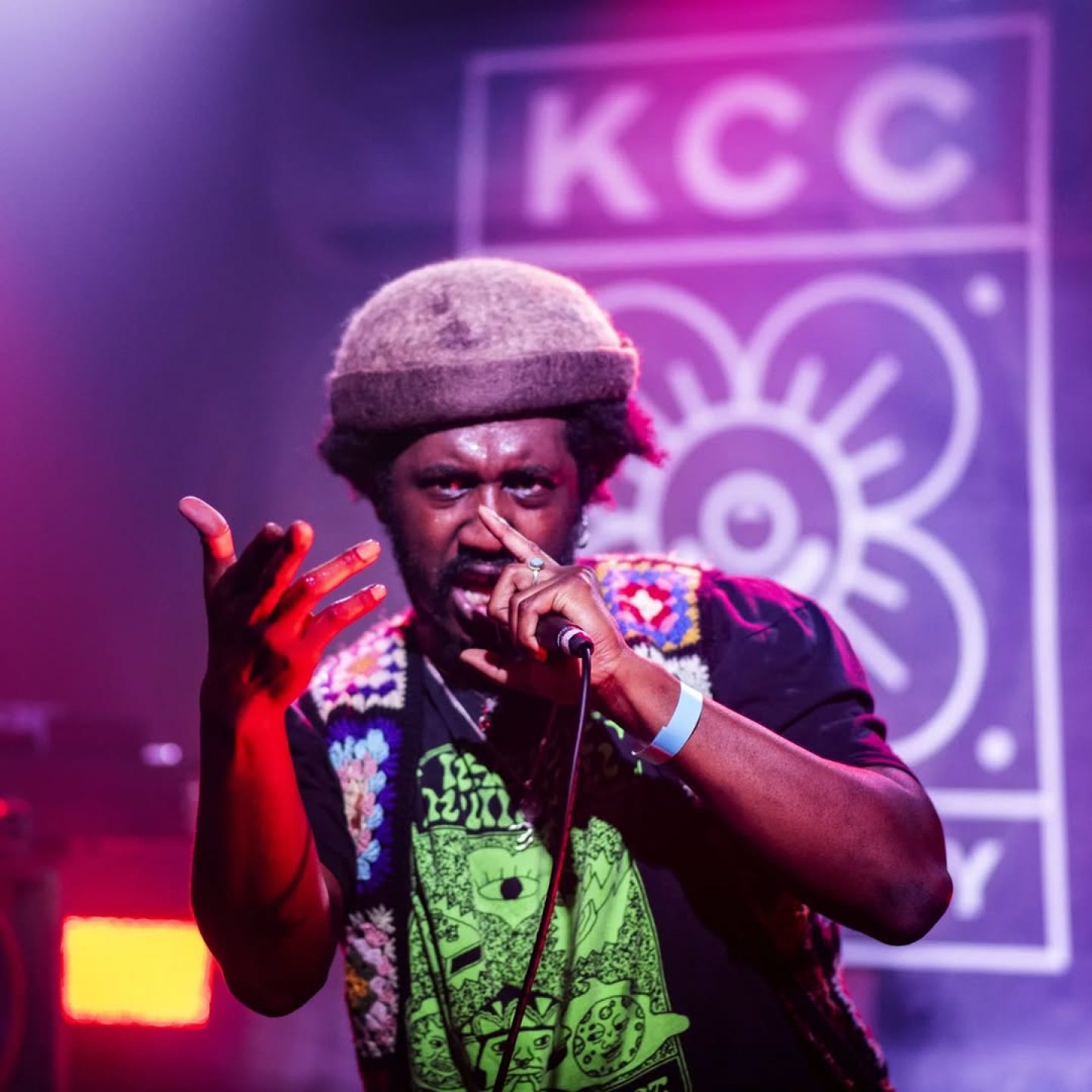 Performer on stage singing into a microphone under purple and pink lights, gesturing toward the audience. A blurred sign with “KCC” and a flower logo is visible in the background, creating a vibrant live music atmosphere.