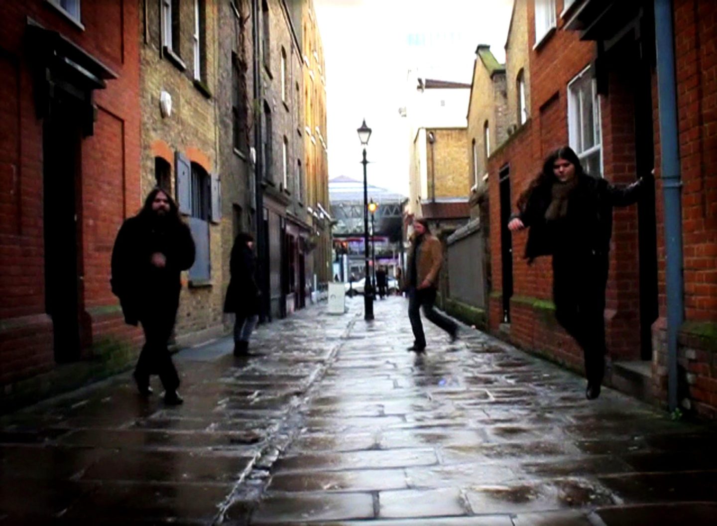 A narrow, wet alleyway lined with brick buildings on both sides. Four people in dark coats are spaced along the street, with one appearing to jump near the right wall. Reflections shimmer on the rain-slick pavement, and a streetlamp stands in the center with brighter buildings visible in the distance.