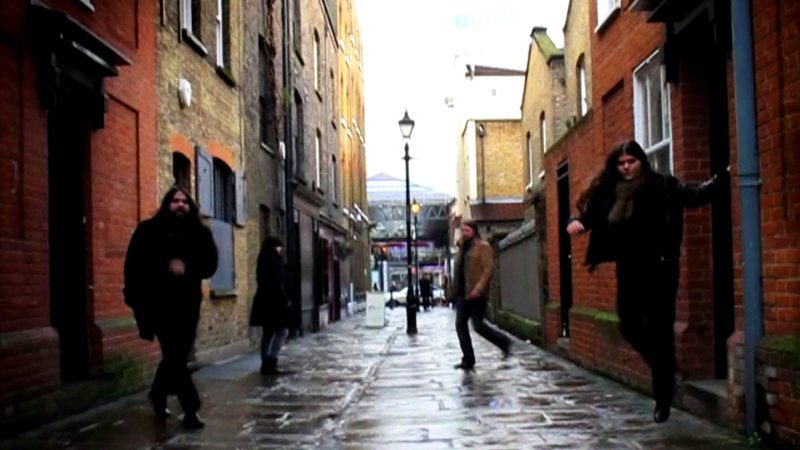 A narrow, wet alleyway lined with brick buildings on both sides. Four people in dark coats are spaced along the street, with one appearing to jump near the right wall. Reflections shimmer on the rain-slick pavement, and a streetlamp stands in the center with brighter buildings visible in the distance.