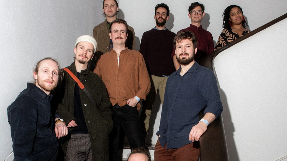 Group portrait of nine musicians arranged on a curved indoor staircase. The group stands on different steps, creating a layered composition. Most wear casual earth-toned or dark clothing, including shirts, jackets and knitwear. White walls and a dark wooden handrail frame the scene, while direct flash photography creates crisp shadows on the walls. The image has the feel of a promotional band photograph, with all members facing the camera and evenly distributed across the staircase.