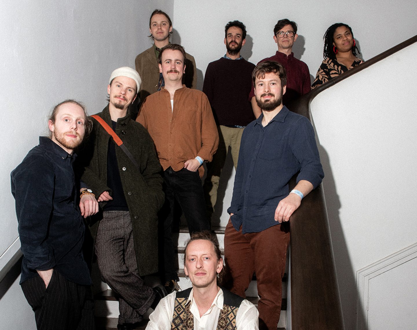 Group portrait of nine musicians arranged on a curved indoor staircase. The group stands on different steps, creating a layered composition. Most wear casual earth-toned or dark clothing, including shirts, jackets and knitwear. White walls and a dark wooden handrail frame the scene, while direct flash photography creates crisp shadows on the walls. The image has the feel of a promotional band photograph, with all members facing the camera and evenly distributed across the staircase.