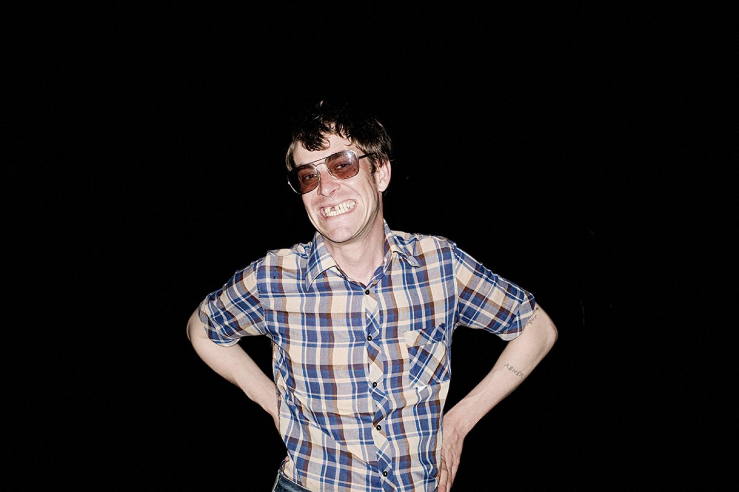 Person wearing tinted sunglasses and a blue plaid shirt poses with hands on hips against a black background, illuminated by direct flash photography.