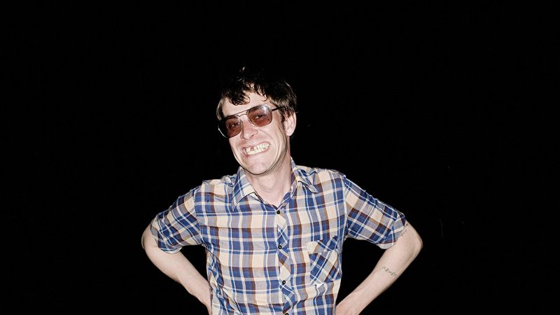 Person wearing tinted sunglasses and a blue plaid shirt poses with hands on hips against a black background, illuminated by direct flash photography.