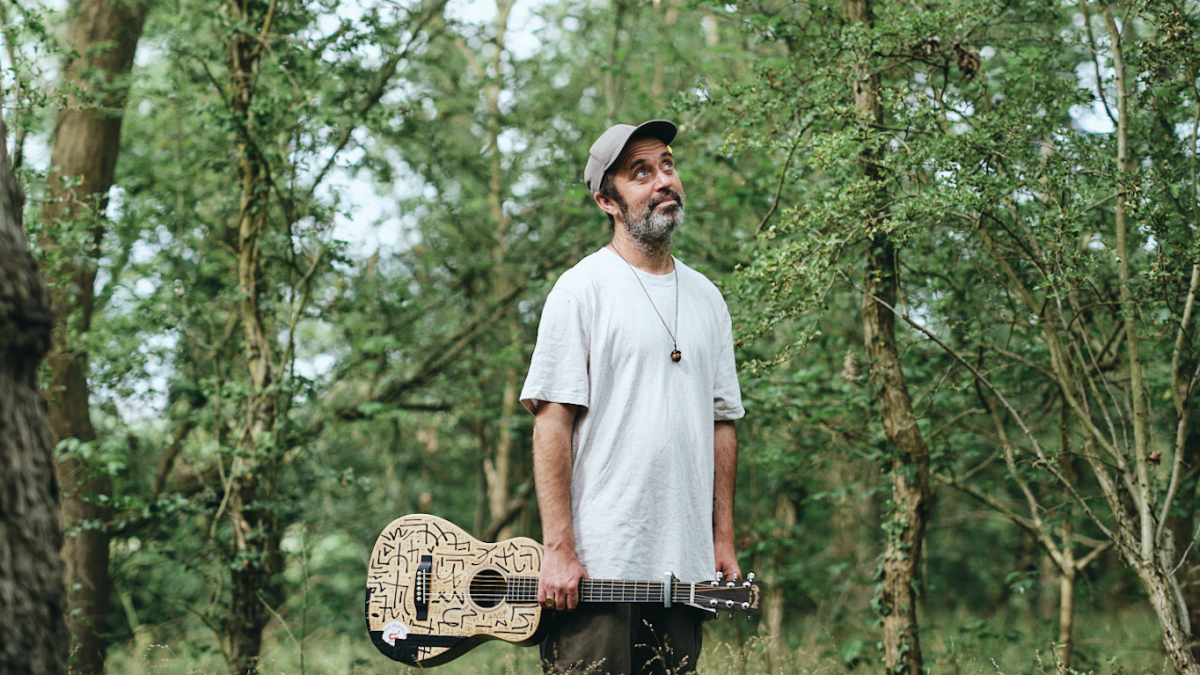 A person standing in a wooded area holding an acoustic guitar and looking up. They are wearing a white t-shirt and a cap.