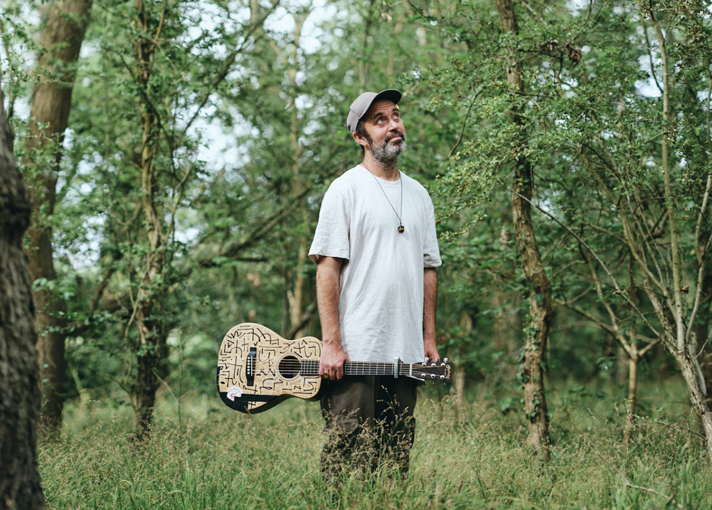 A person standing in a wooded area holding an acoustic guitar and looking up. They are wearing a white t-shirt and a cap.