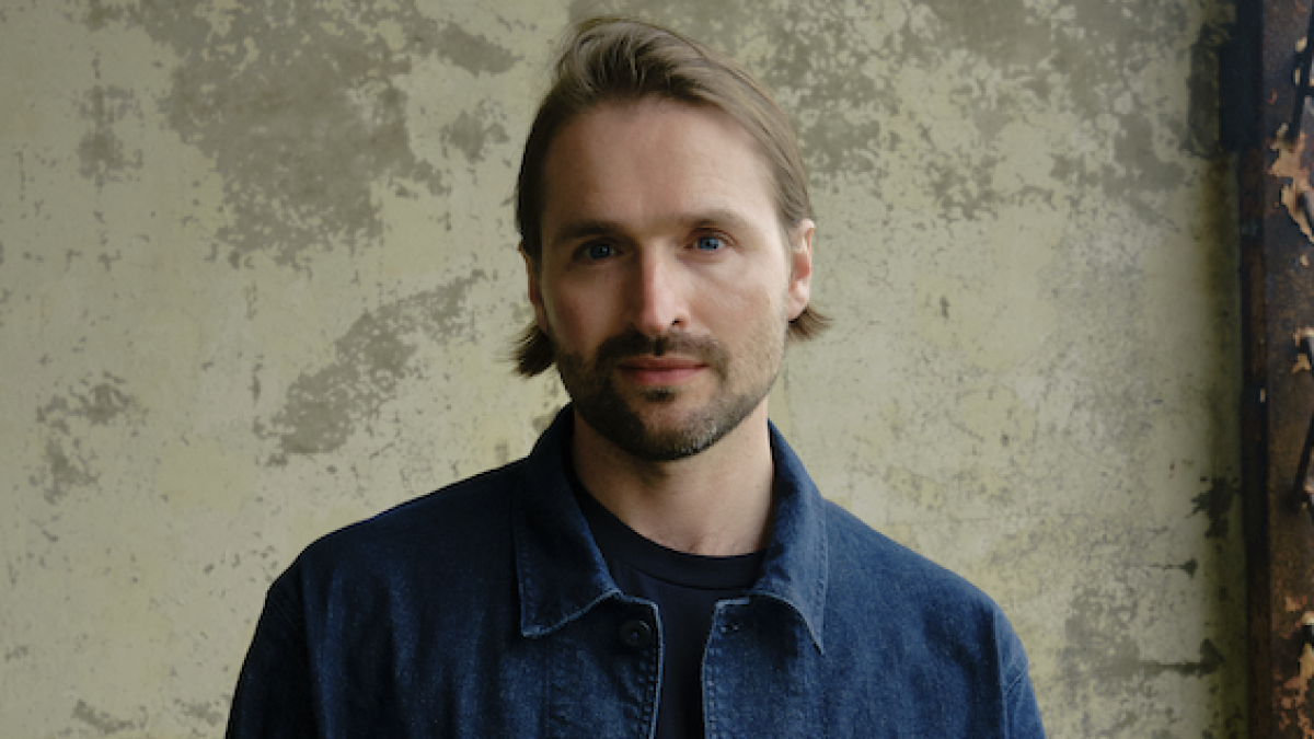 A person with medium-length light brown hair swept back and a short beard looks directly at the camera with a neutral expression. They are wearing a dark navy jacket over a black shirt and standing in front of a weathered beige wall with a textured, peeling finish. Soft natural light illuminates their face, creating a calm, understated portrait.