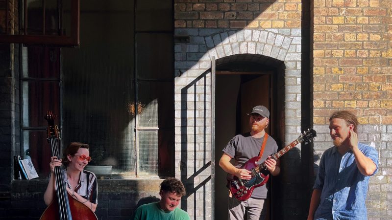 Tom Moore Quartet Four musicians with a double bass, drum, guitar and keyboard stand in a sunlit cobbled courtyard outside a brick building, with a black cat sitting in the foreground.