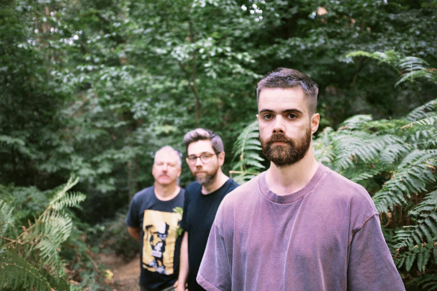 Three men walking through a lush green forest. A bearded man in a mauve T-shirt is in sharp focus in the foreground, while two men follow behind him slightly out of focus. Ferns and dense woodland surround them.