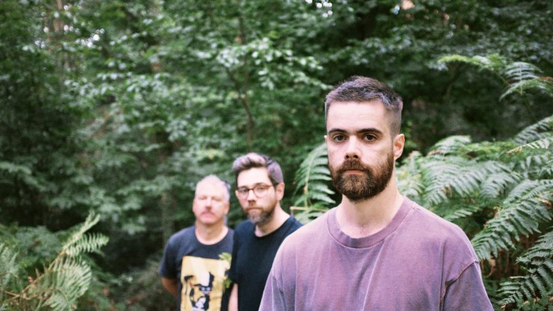Three men walking through a lush green forest. A bearded man in a mauve T-shirt is in sharp focus in the foreground, while two men follow behind him slightly out of focus. Ferns and dense woodland surround them.