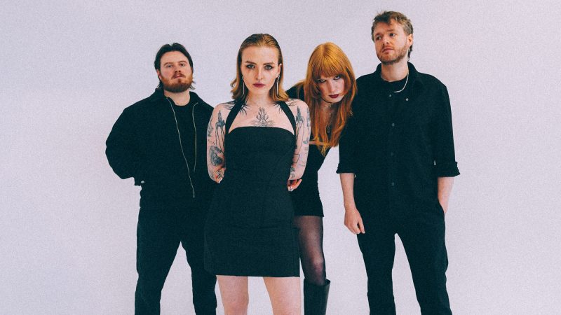 Four band members dressed in black pose against a plain light background, with one tattooed person standing in front and three others arranged behi