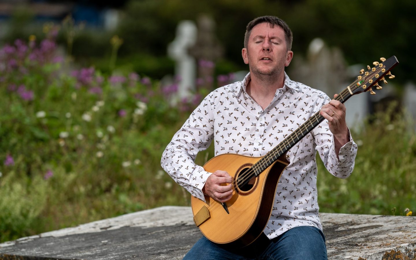 A portrait photo by Eddie Cavanagh of folk musician Daoirí Farrell playing an Irish bouzouki outdoors while seated on a stone ledge