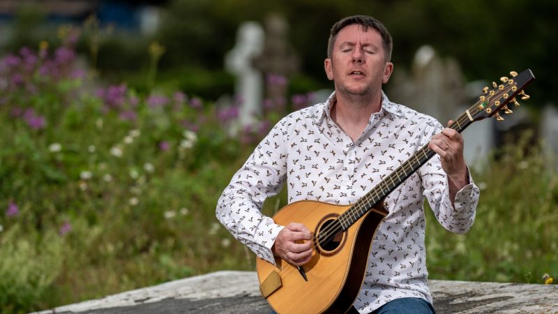 A portrait photo by Eddie Cavanagh of folk musician Daoirí Farrell playing an Irish bouzouki outdoors while seated on a stone ledge
