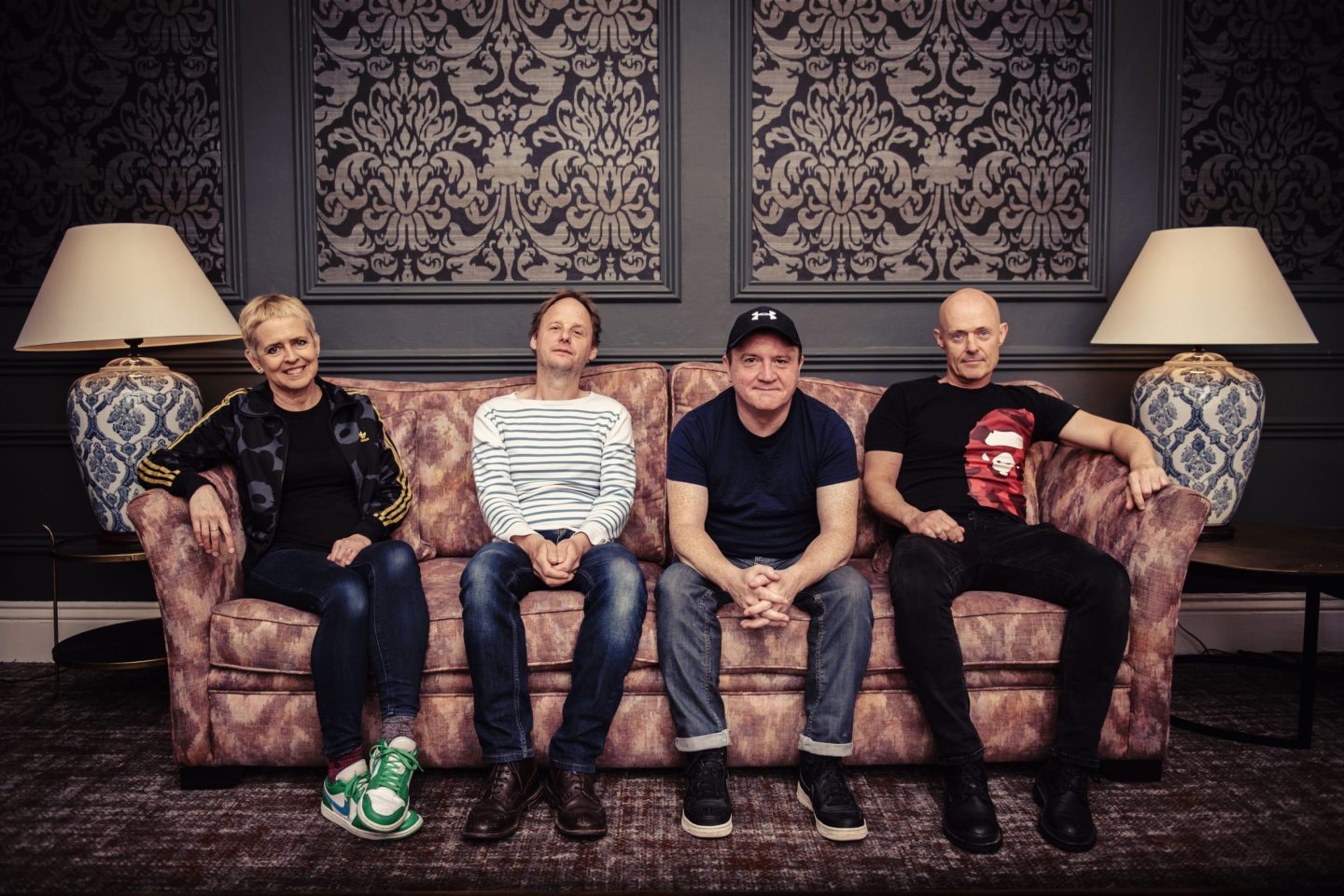 A band profile photo by Eddie Kavanagh showing the four members of Flook seated together on a patterned sofa against an ornate wallpapered wall framed by two decorative lamps