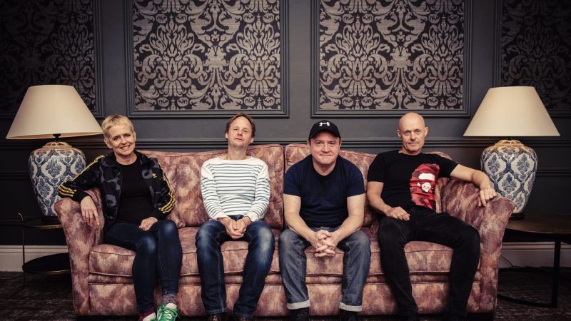 A band profile photo by Eddie Kavanagh showing the four members of Flook seated together on a patterned sofa against an ornate wallpapered wall framed by two decorative lamps