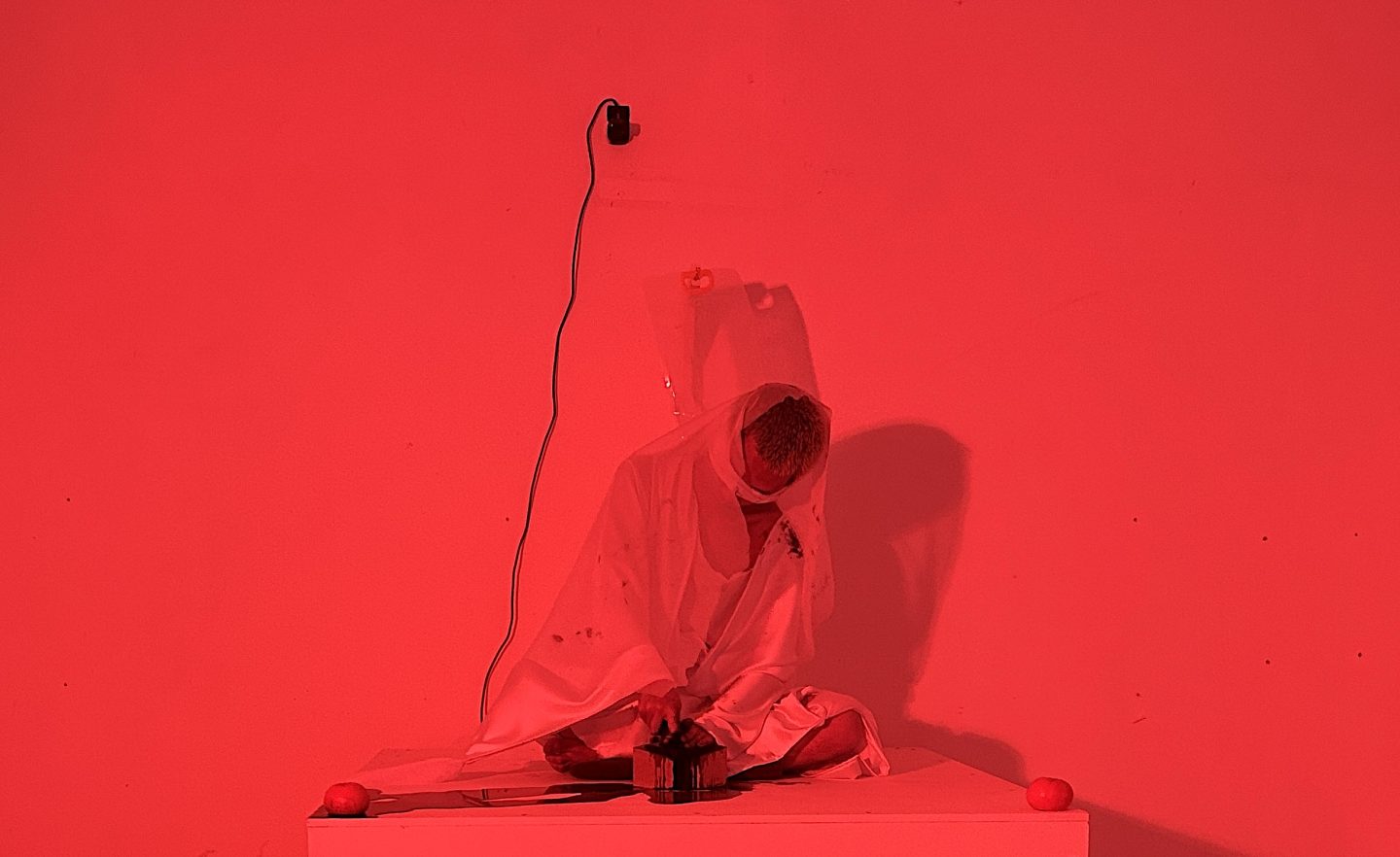An artistic performance illuminated by vivid red light, featuring a figure draped in white fabric seated cross-legged atop a low white platform