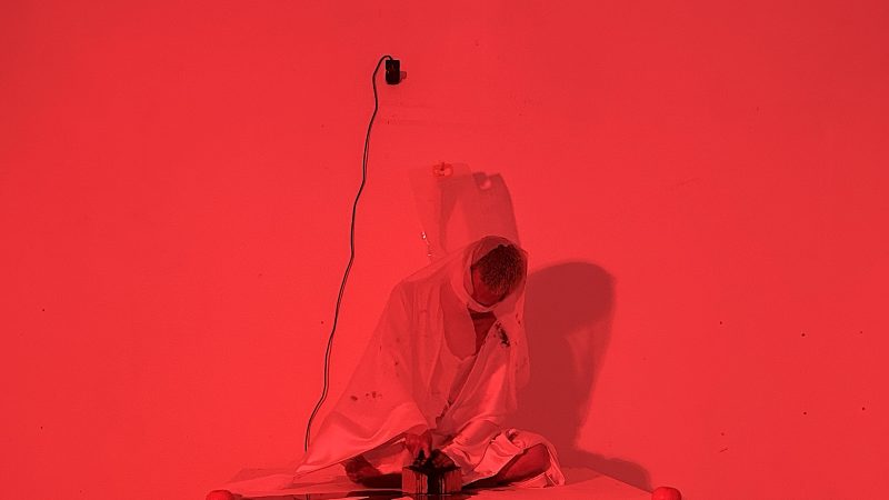 An artistic performance illuminated by vivid red light, featuring a figure draped in white fabric seated cross-legged atop a low white platform
