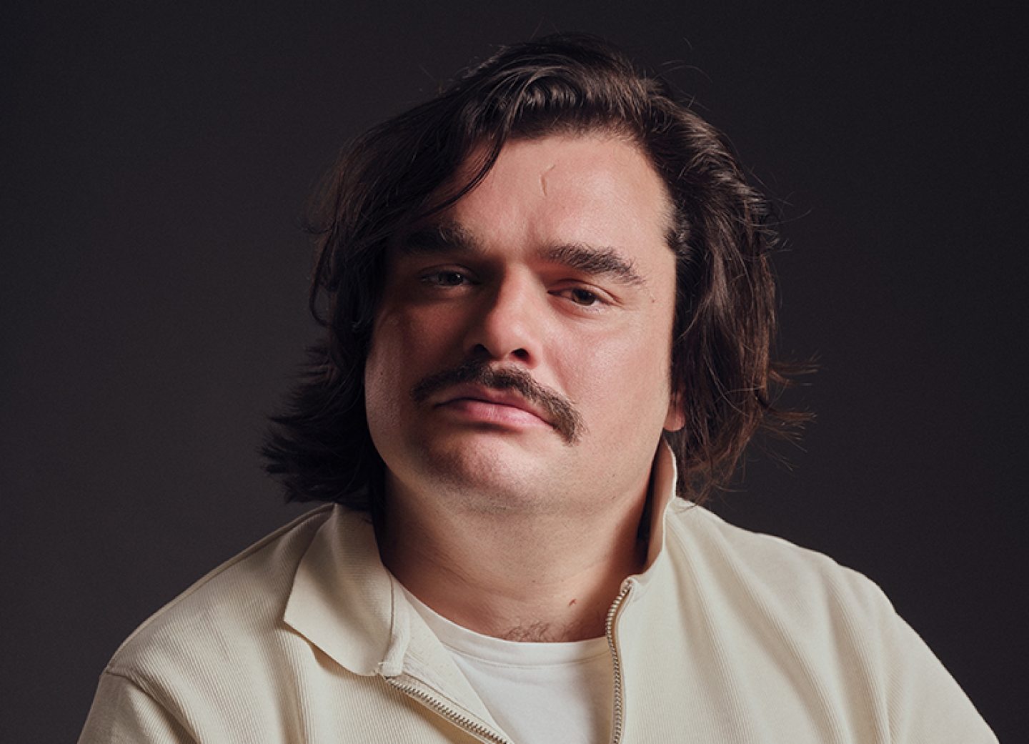 Studio portrait of a Red Richardson, with shoulder-length dark hair and a thick moustache, wearing a cream zip-up jacket over a white shirt, looking toward the camera against a dark, softly lit background.
