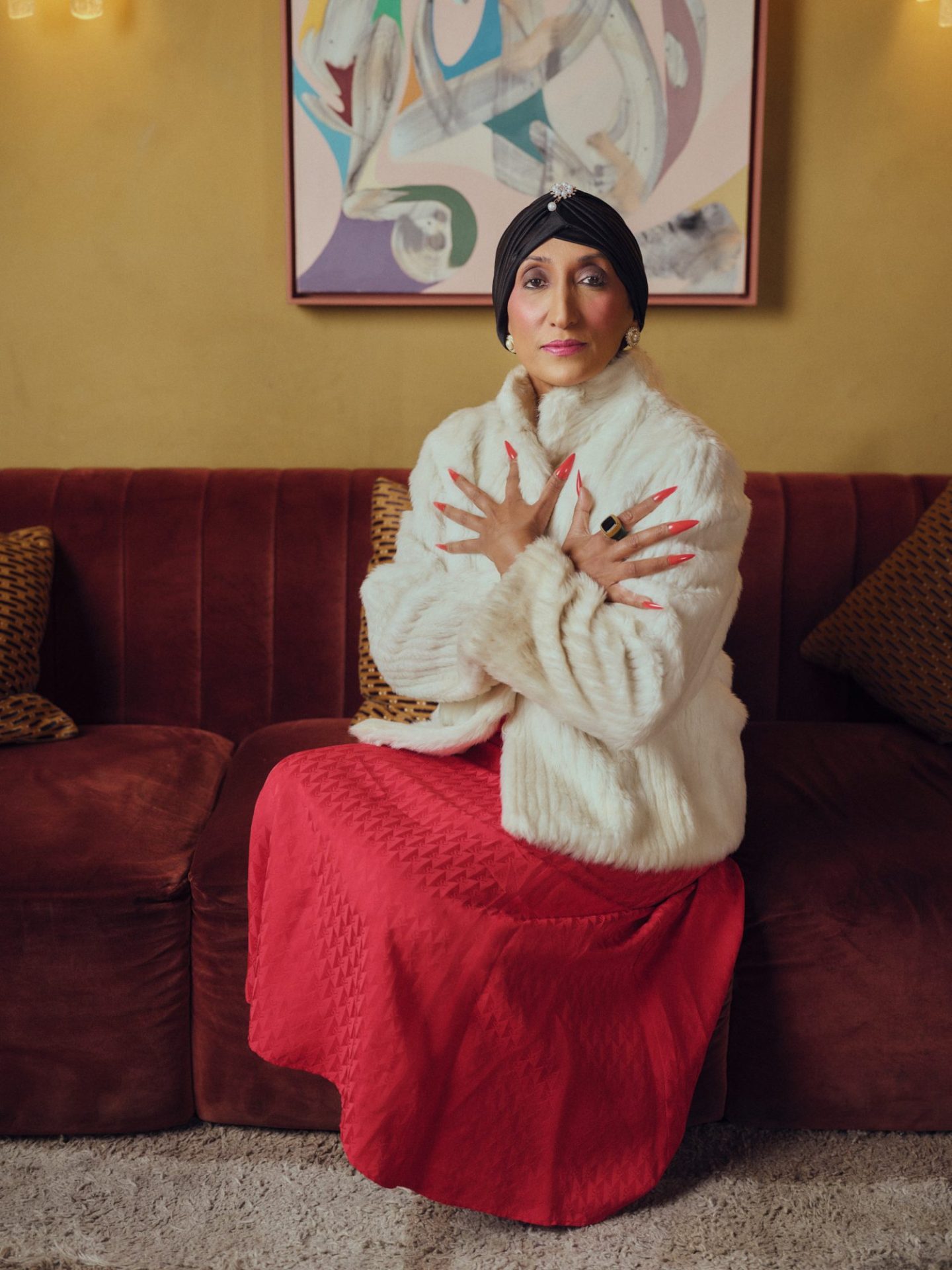 Shazia Mirza, A person in a white faux-fur coat and red skirt posing on a burgundy velvet sofa beneath an abstract painting in a warm, stylish interior.
