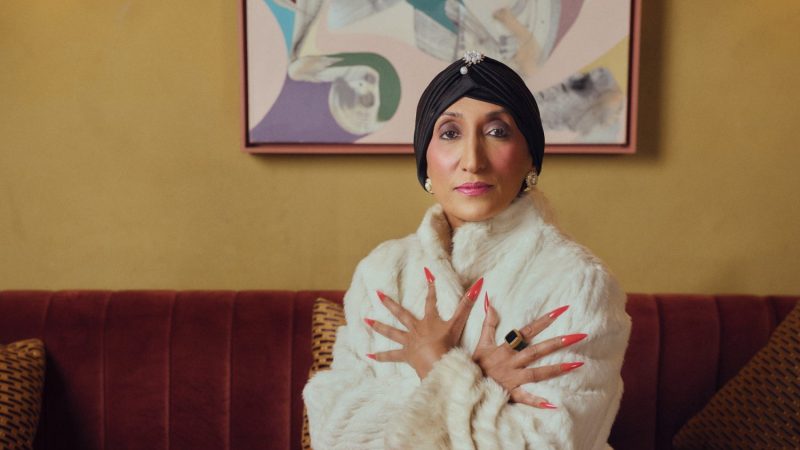 Shazia Mirza, A person in a white faux-fur coat and red skirt posing on a burgundy velvet sofa beneath an abstract painting in a warm, stylish interior.