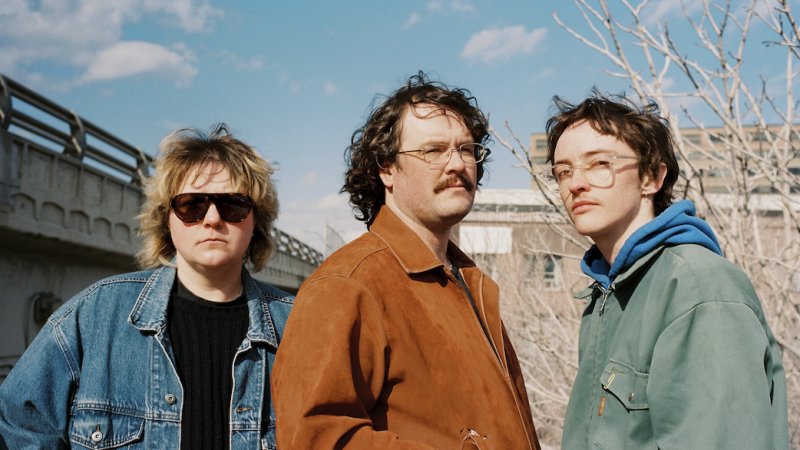A band profile photo featuring three members outdoors in natural sunlight, set against an urban industrial backdrop with concrete structures and bare trees under a clear blue sky.
