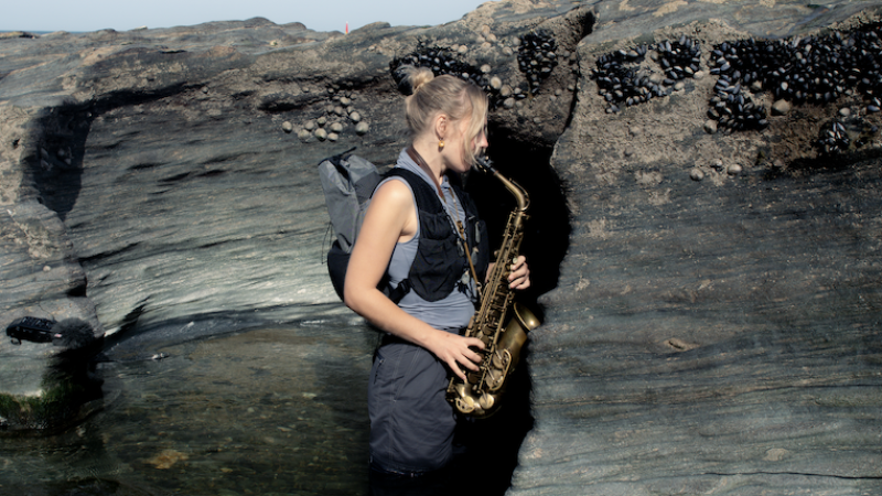 Laura Misch is standing in a rocky tidal pool, playing a saxophone beside layered coastal rock formations.