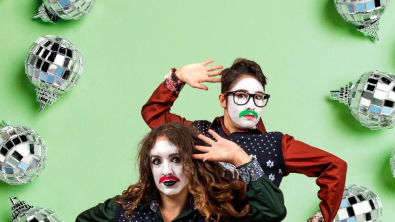Two performers, Sh!t Theatre, in festive sweaters, shorts, and face paint pose against a green background surrounded by mirrored disco-ball ornaments.