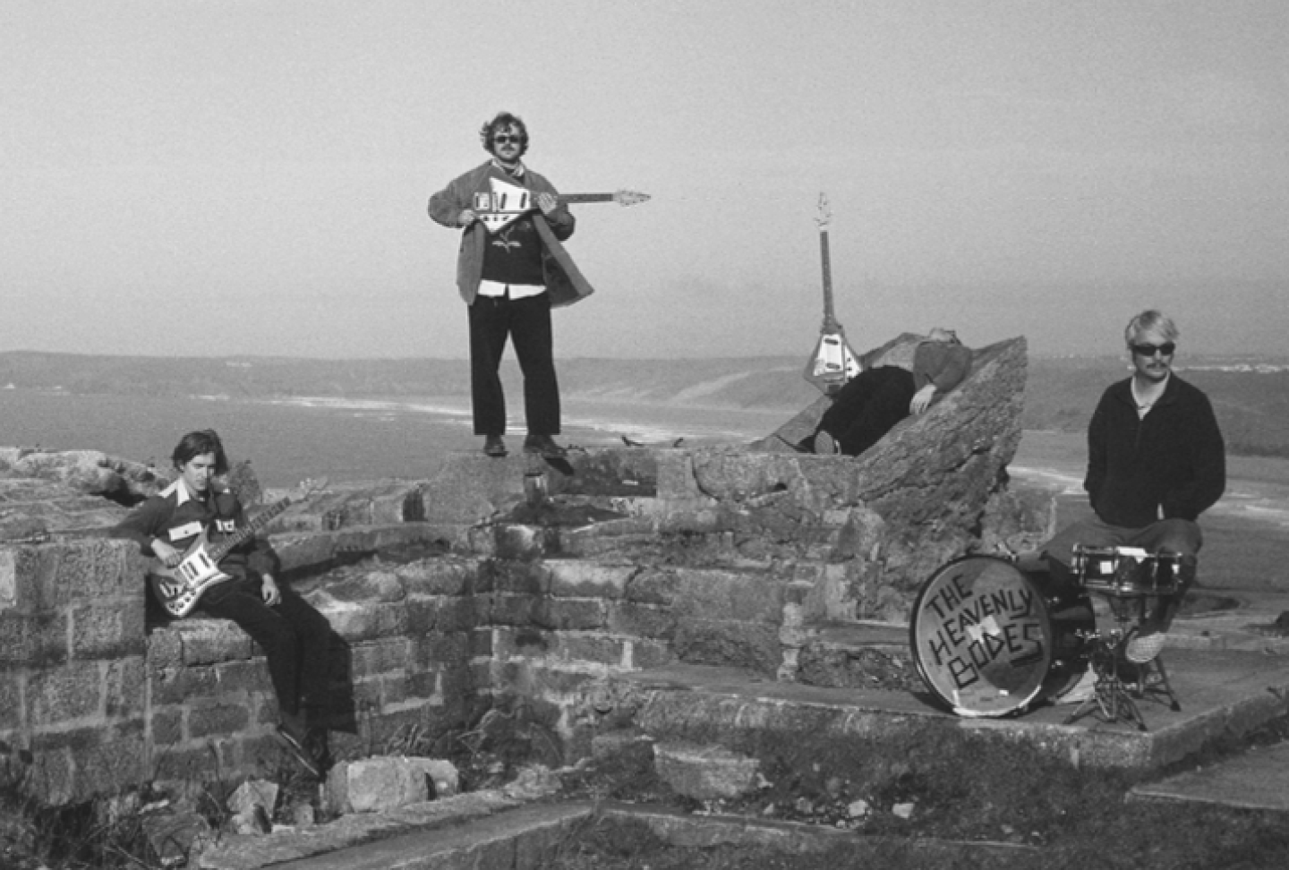 A black-and-white promotional photo of the band The Heavenly Bodes posing with their instruments outdoors on stone ruins overlooking a coastline.