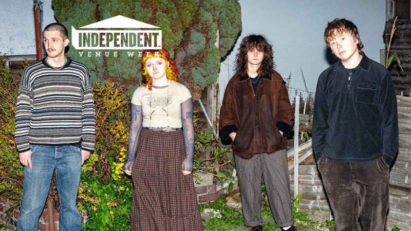 Four band members The New Cut standing in a garden at night, photographed with flash against shrubs and a large tree, with an Independent Venue Week logo in the corner.