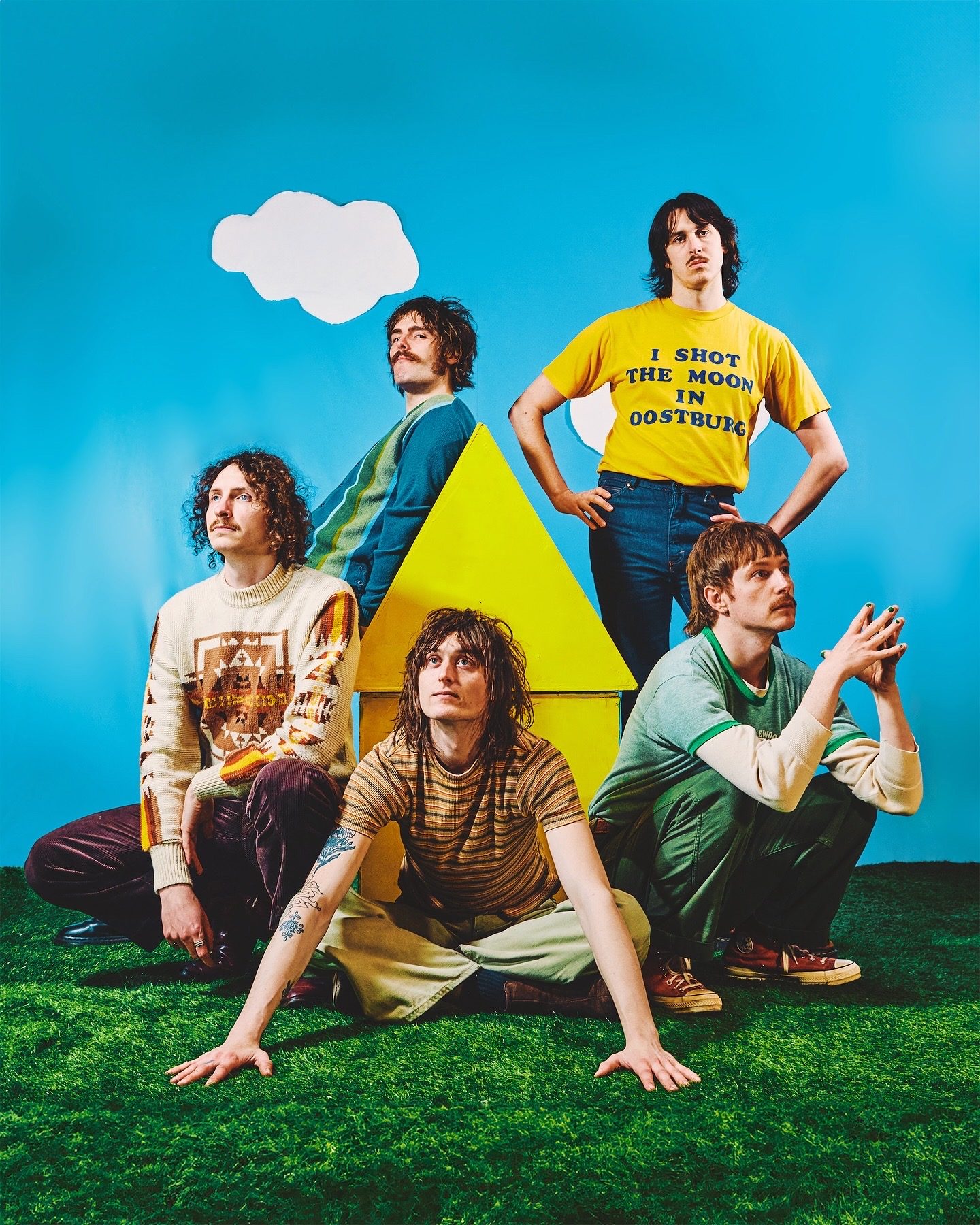 A colorful studio band portrait of five musicians posing around a yellow house prop on artificial grass against a bright blue backdrop with paper cutout clouds