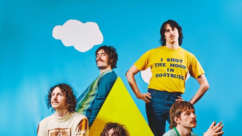 A colorful studio band portrait of five musicians posing around a yellow house prop on artificial grass against a bright blue backdrop with paper cutout clouds
